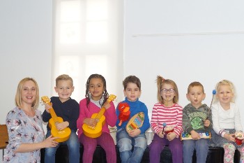 sechs Kinder sitzen nebeneinander, jedes hat ein kleines Musikinstrument in der Hand. Links steht die Lehrerin