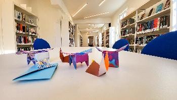 fünf Origami-Figuren auf einem weißen Tisch , links und rechts sind Regale voller Bücher zu sehen.