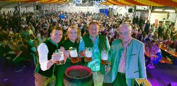 drei Herren und eine Dame mit gefüllten Bierkrügen in der Hand stehen auf der Bühne, im HIntergrund volles Festzelt