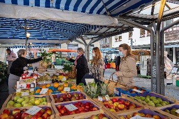 Marktstand mit Auslage von Obst und Gemüse, einkaufende Frauen werden am Stand bedient.