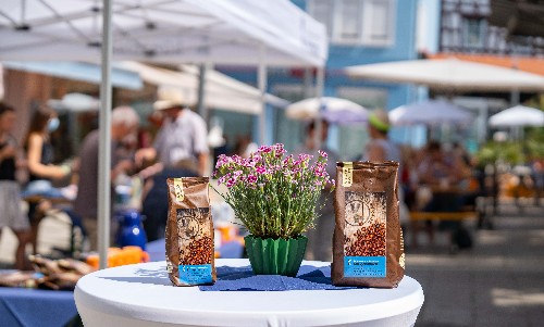 Stehtisch mit 2 Kaffeepackunegn und einem blühenden Blumentopf, im HIntergrund Menschen unter Sonnenschirmen